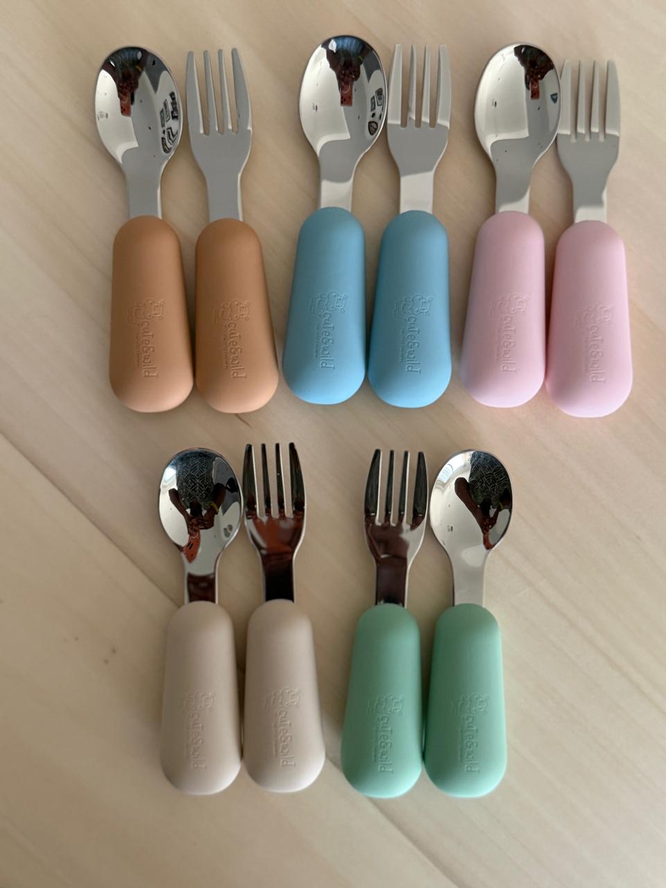 A variety of cute&wild baby utensils on a wooden board, including stainless steel forks and spoons with colorful silicone handles and a 4-pack of flexible silicone spoons.